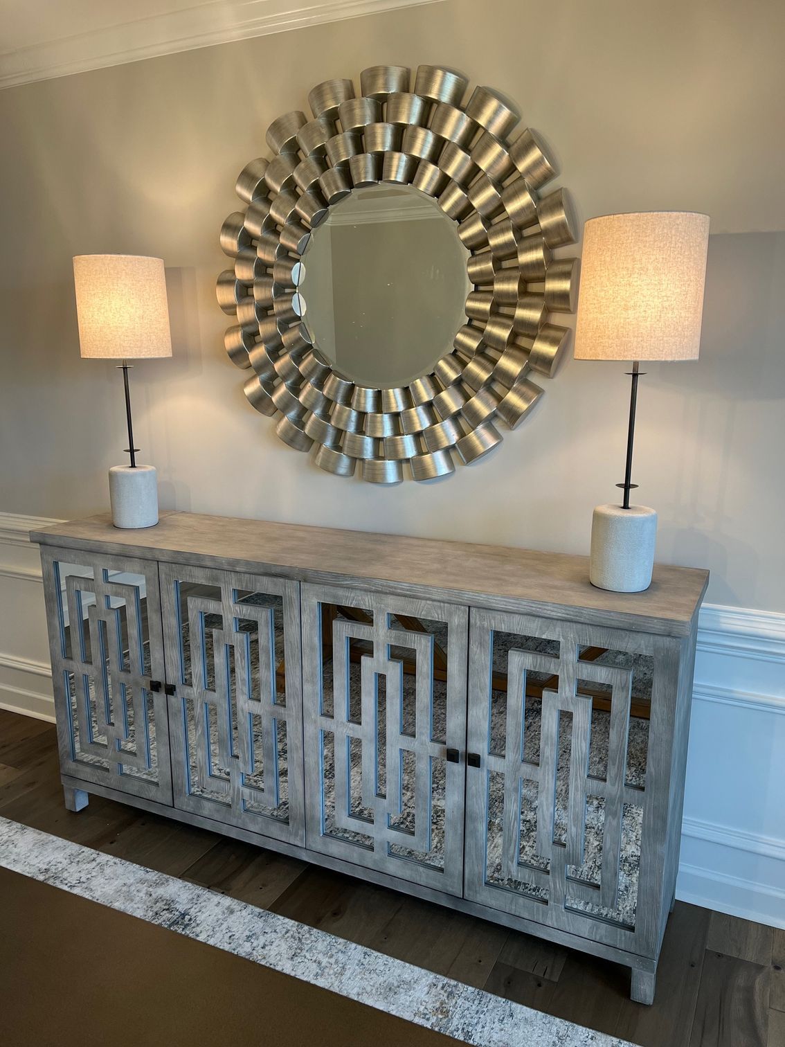 A gray cabinet with openwork doors is below a sunburst mirror and two lamps.