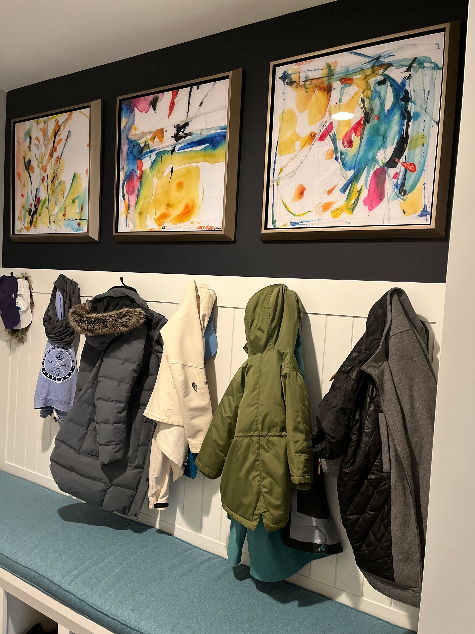 Hallway with artwork above coat hooks and a bench. Coats hang from hooks; art has colorful, abstract designs.