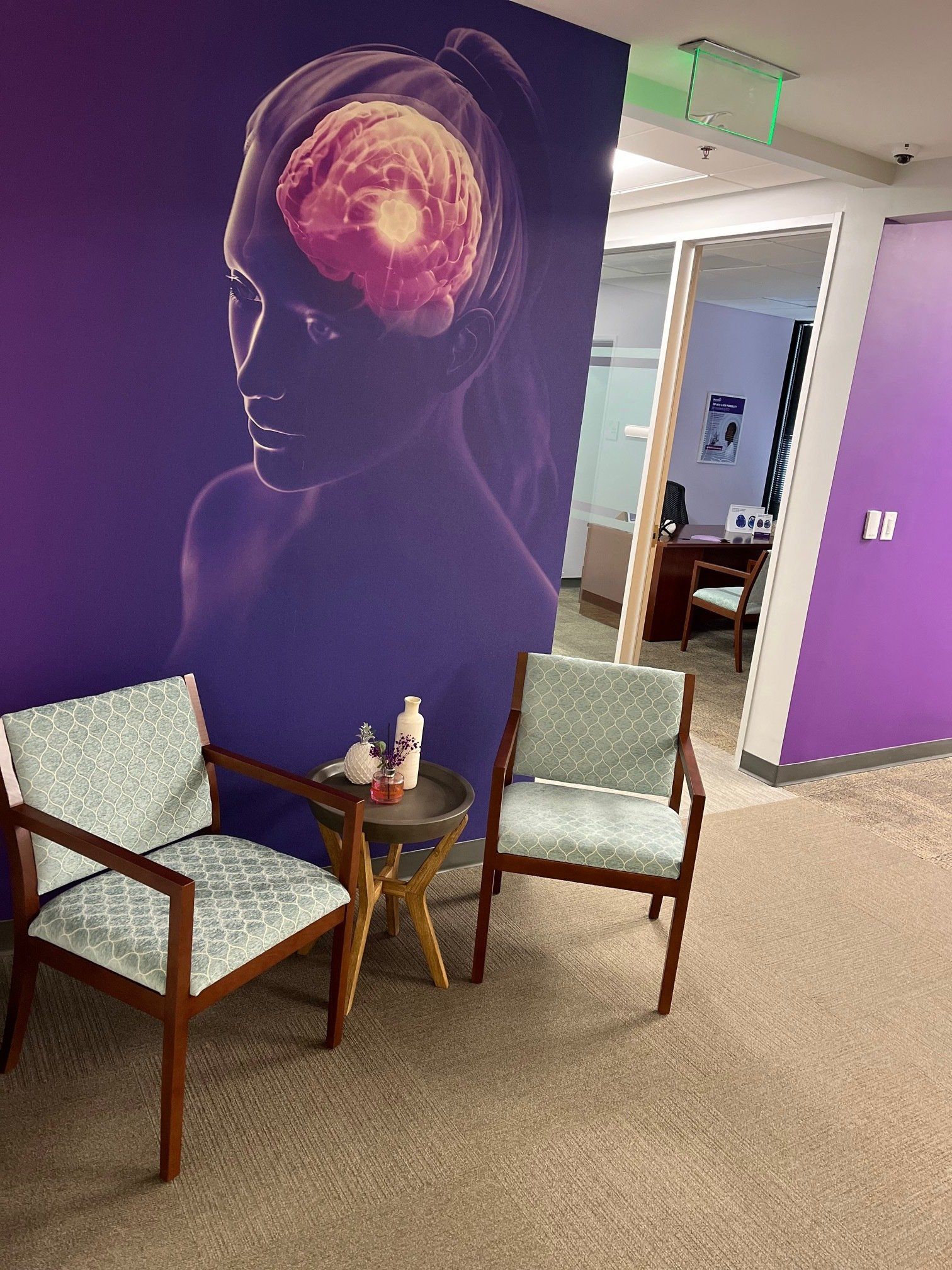 Two chairs in a waiting room with a brain graphic, purple wall, and view of an office.