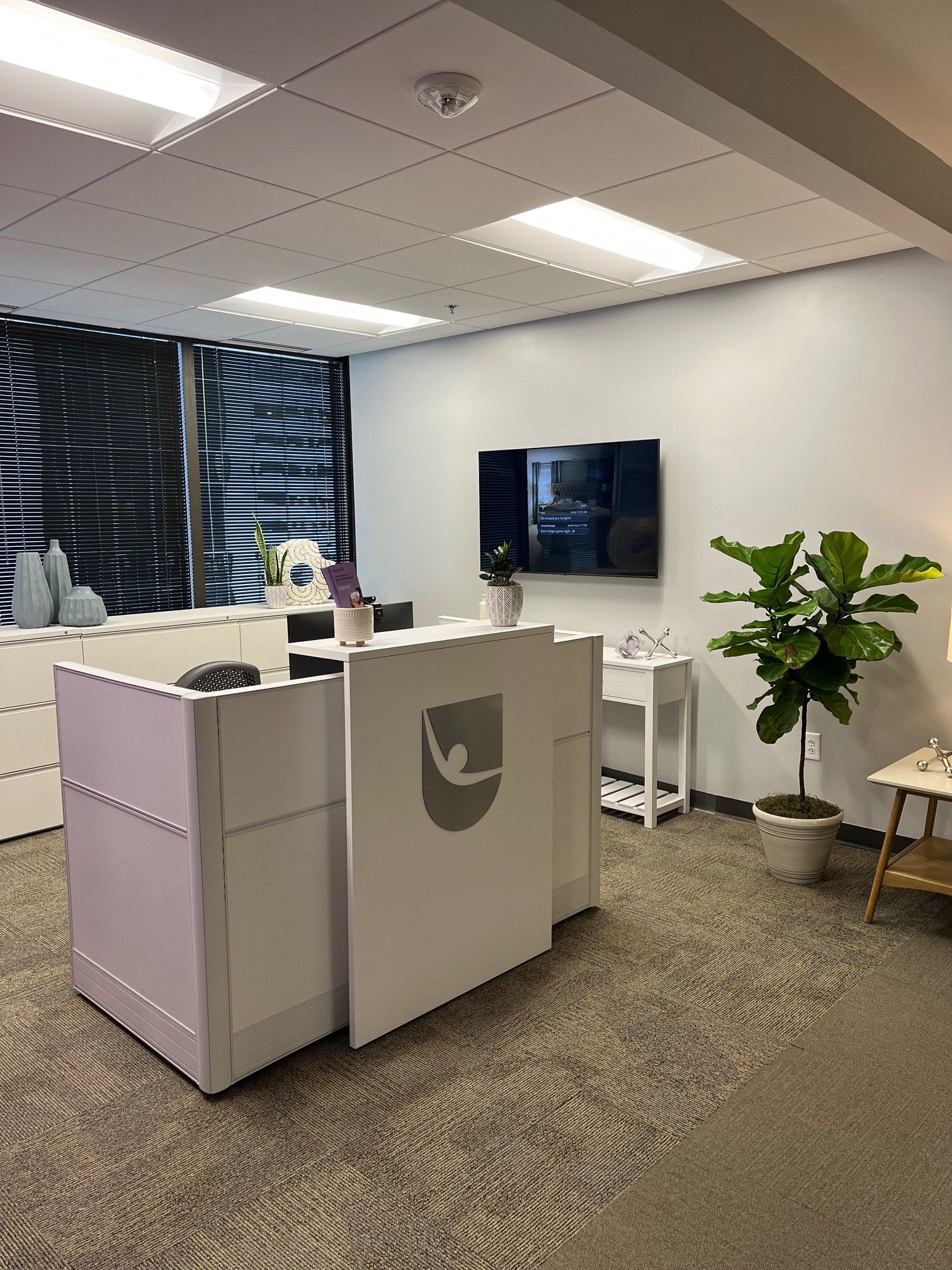 White reception desk with logo in a bright office; a TV and plant are visible.