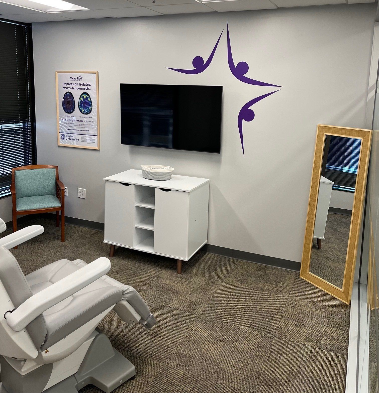 A medical room with a therapy chair, TV, cabinets, chair, mirror and wall decor.
