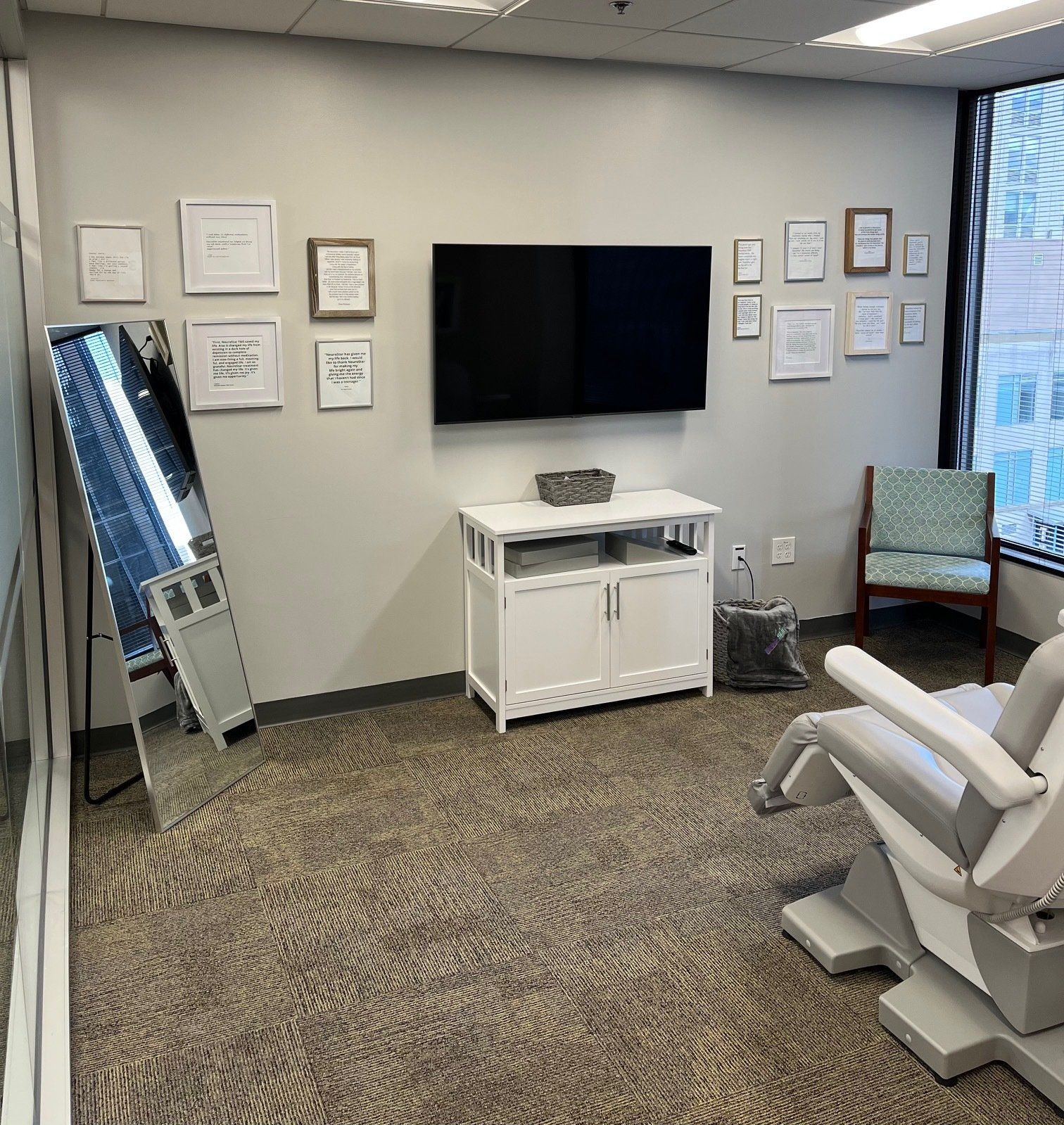 Room with dental chair, TV, framed art, white cabinet, and tall mirror.