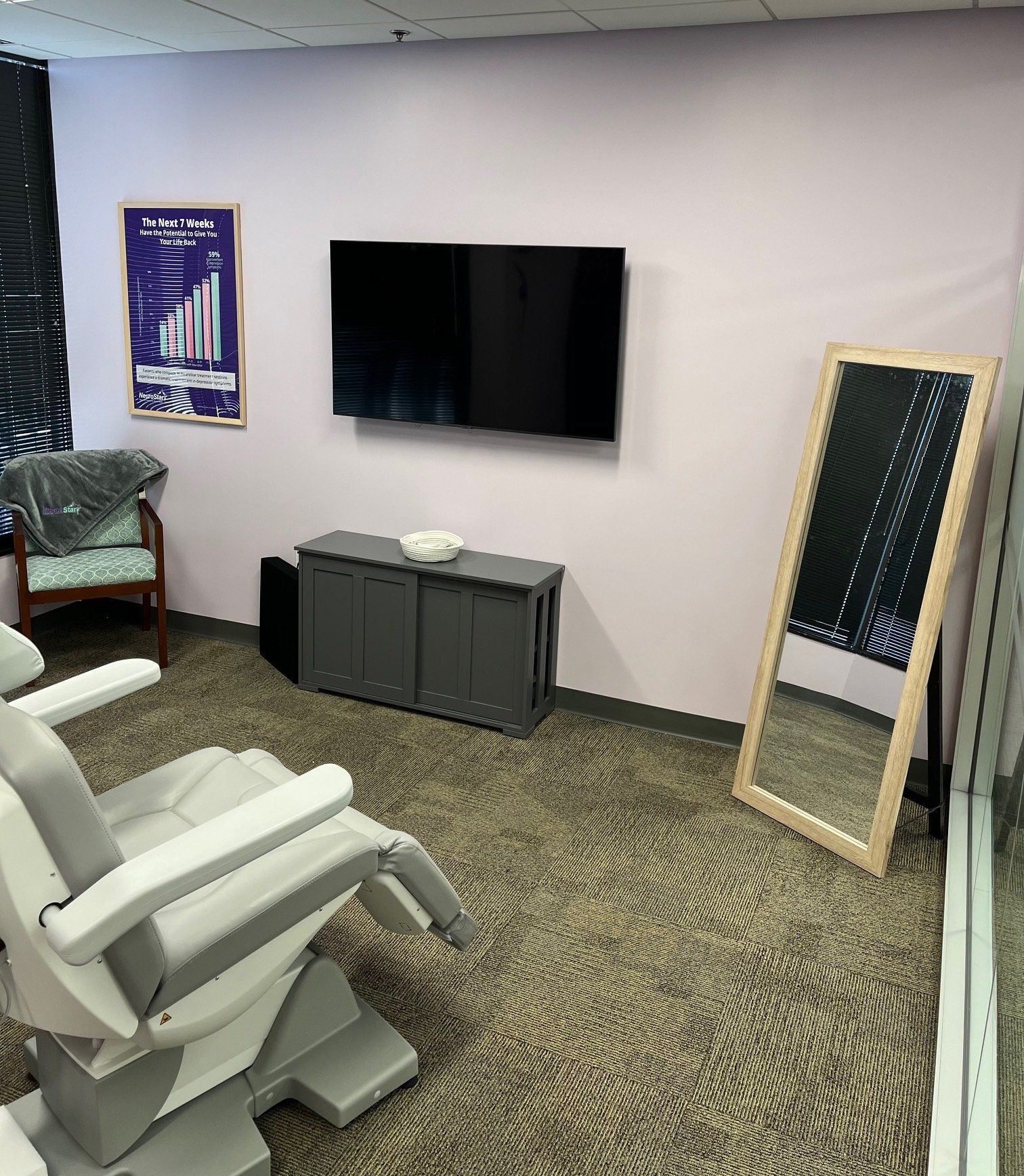 A medical room with a chair, TV, mirror, and cabinet against a pink wall; chair is grey and white.