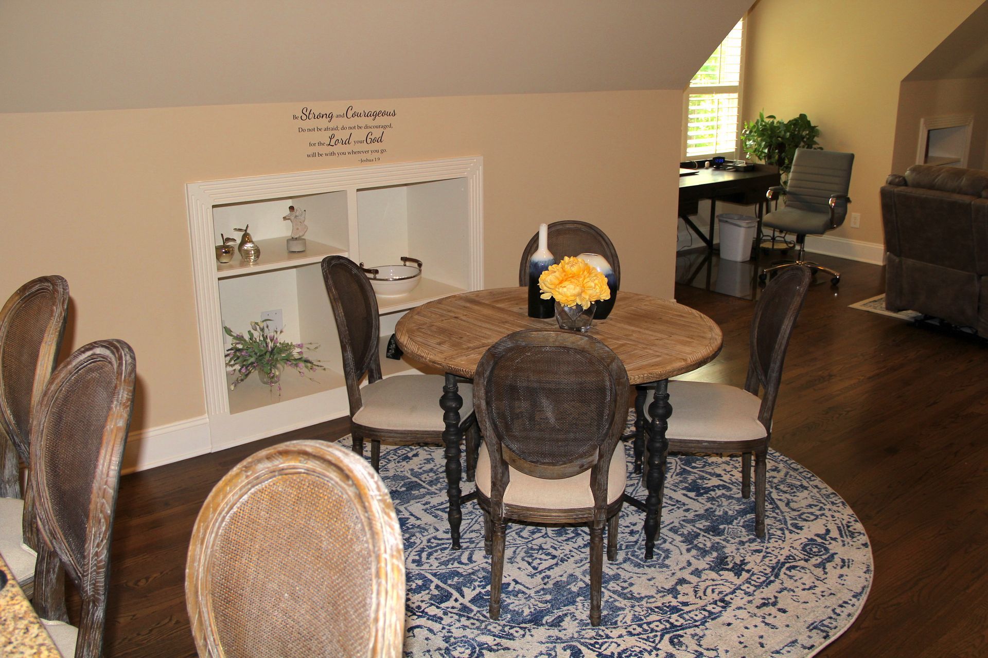 Dining area with round table, four chairs on a blue rug. Niche with shelves.