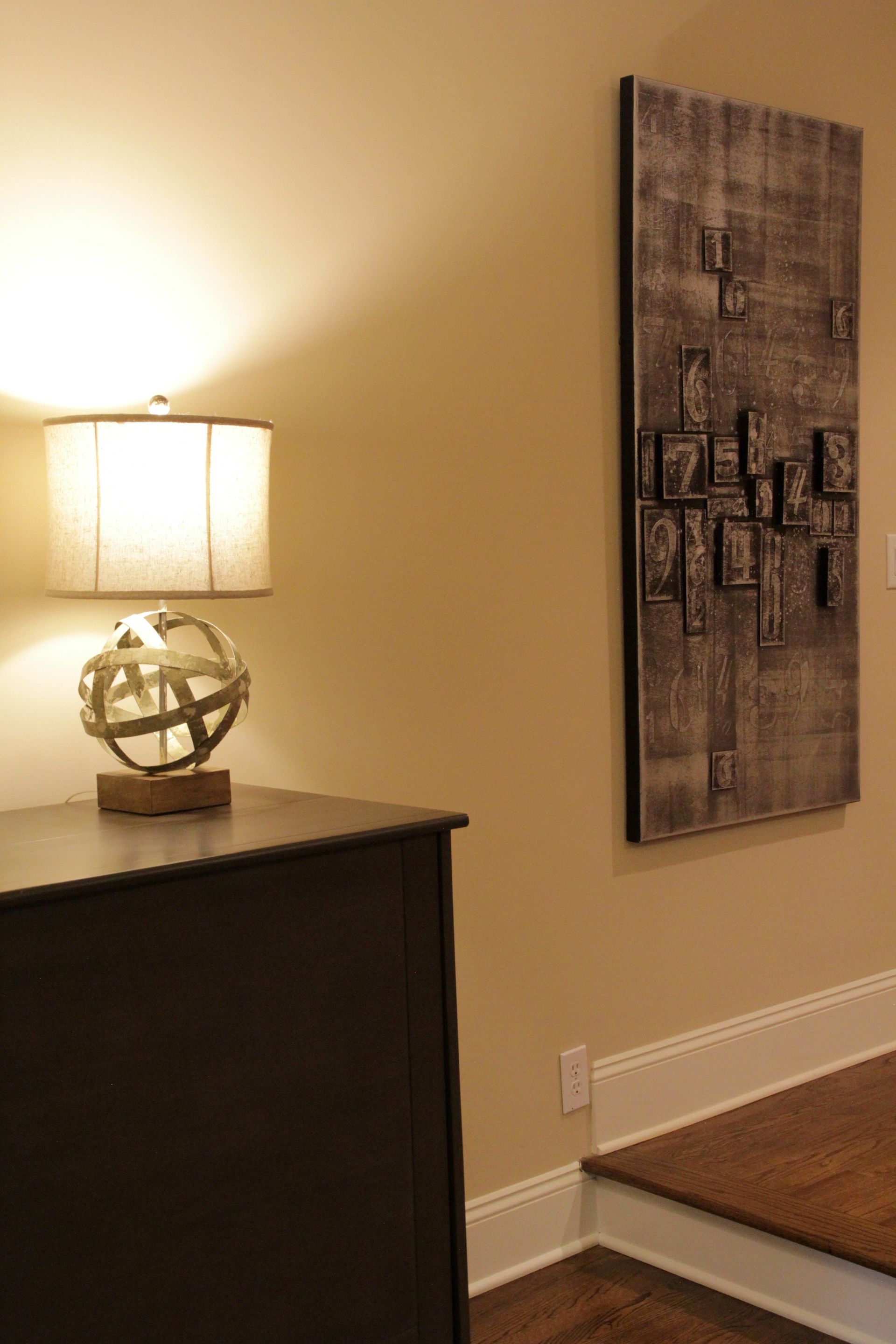 A lamp on a dark table next to a textured wall art on a beige wall.