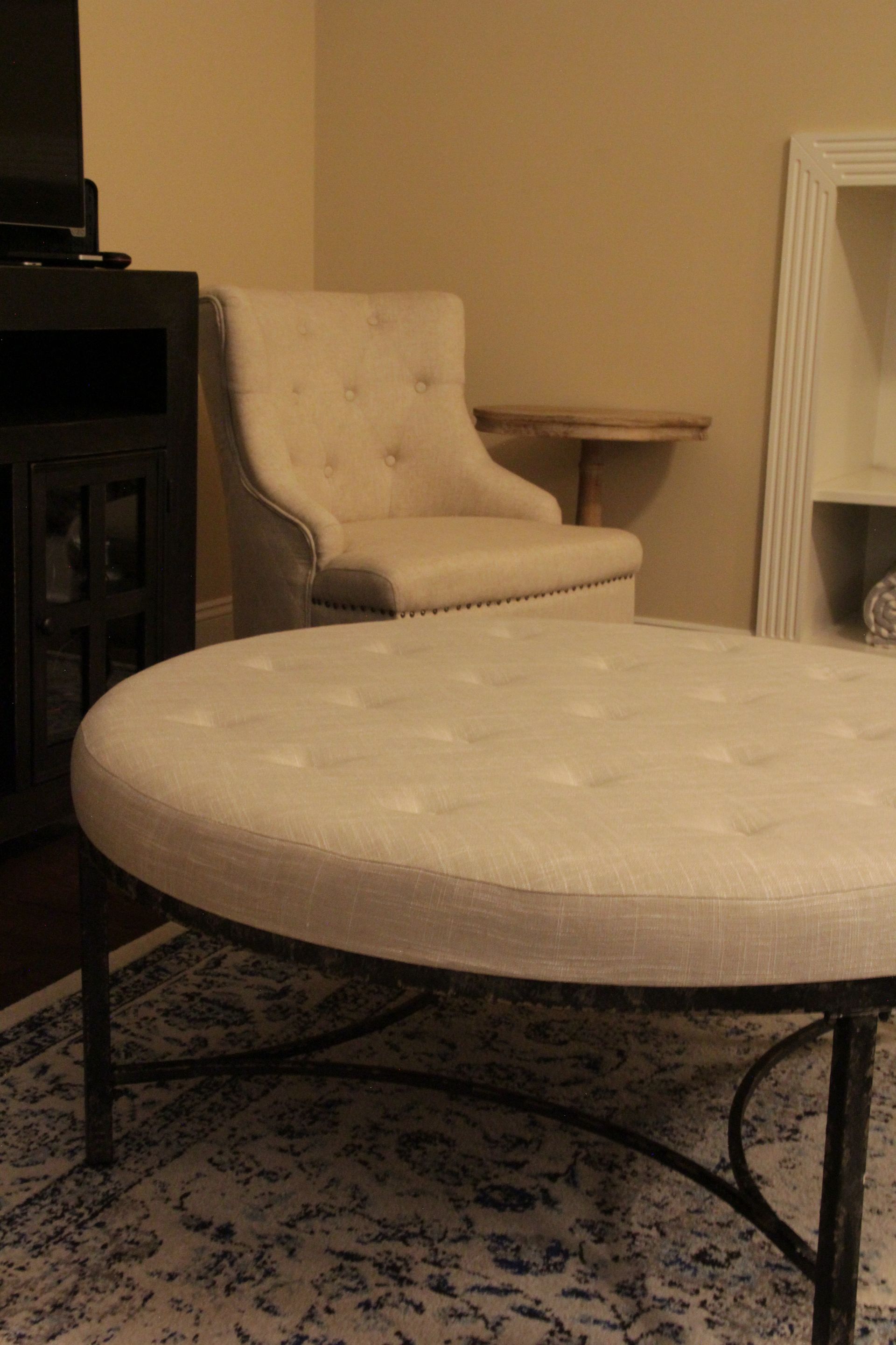 Round ottoman with tufted cream top and wrought iron base, sitting on patterned rug, chair in background.
