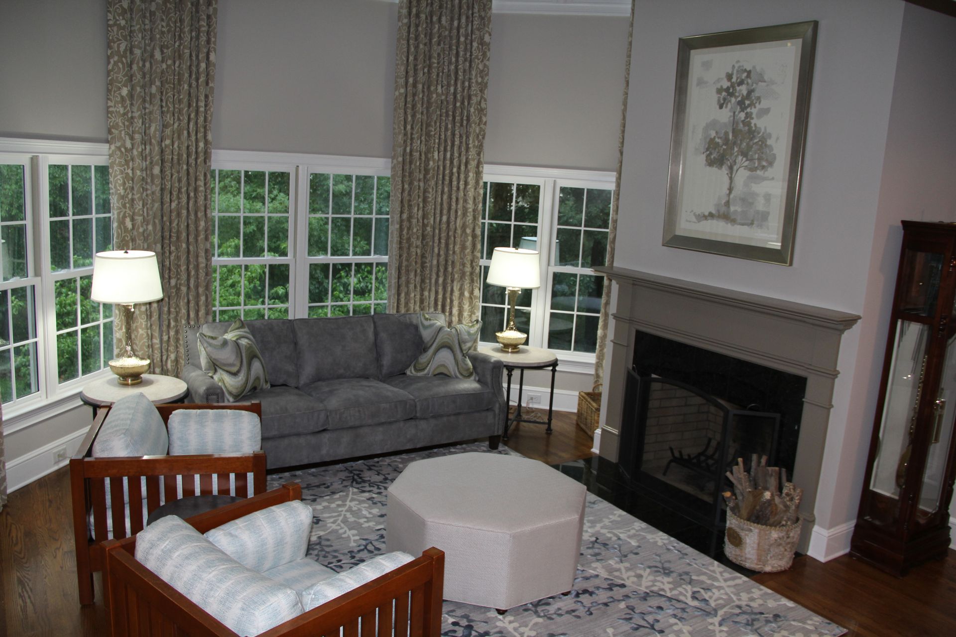 Cozy living room with a gray sofa, two armchairs, a rug, a fireplace, and large windows with patterned curtains.