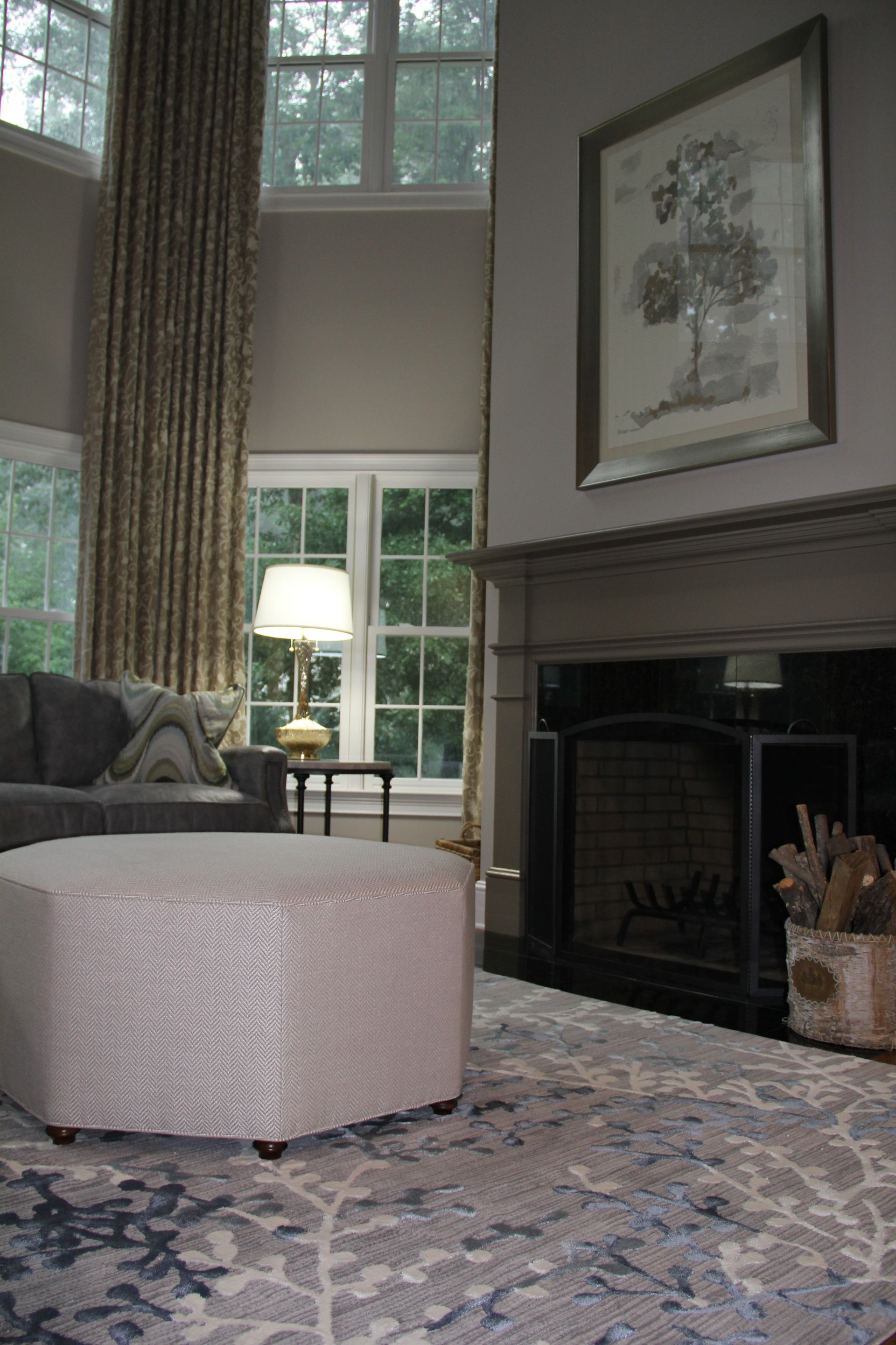 Living room: gray sofa, patterned ottoman, fireplace with art, floral rug, windows, and drapes.
