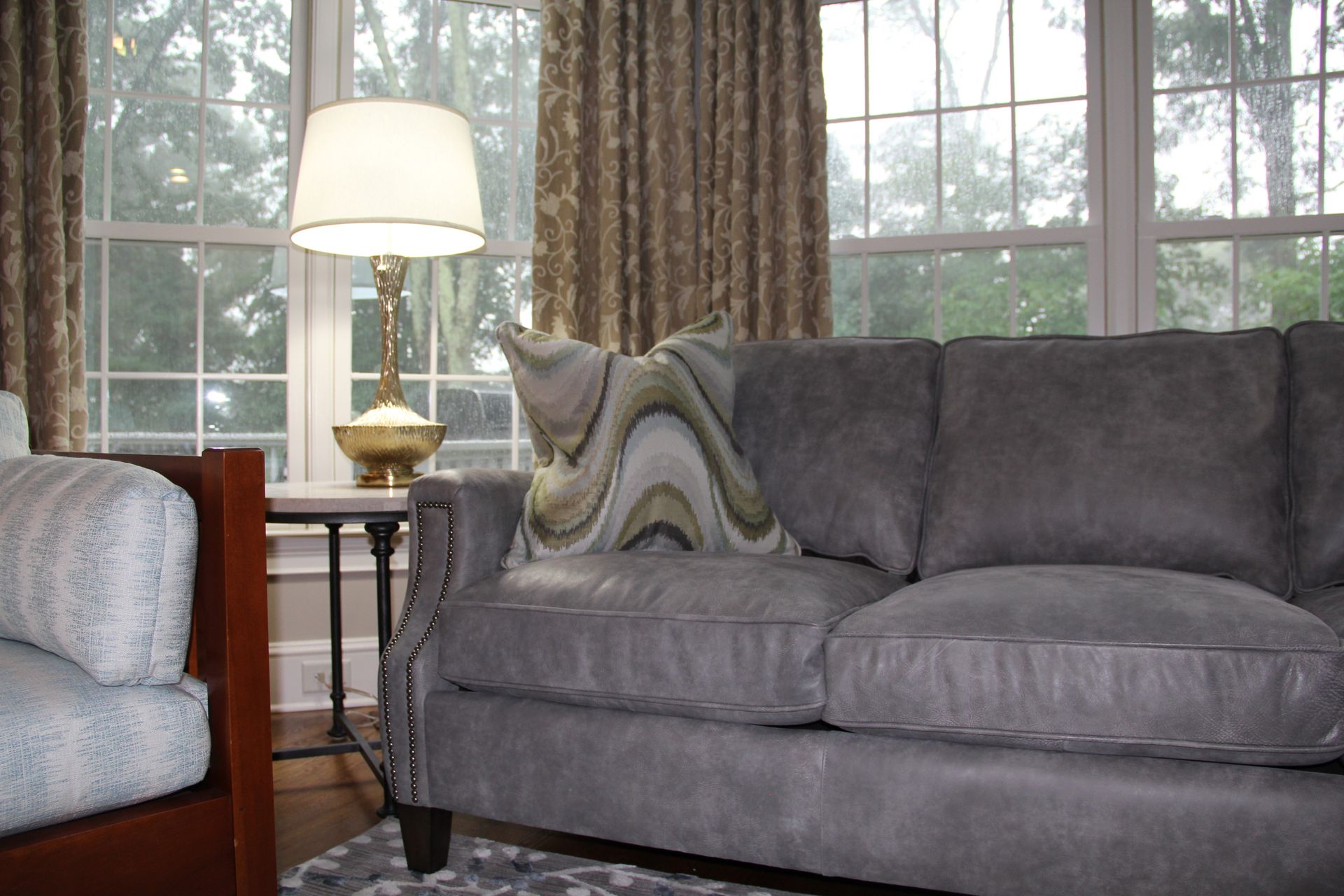 Gray sofa, accent pillow, lamp, and patterned curtains in a living room with windows overlooking trees.