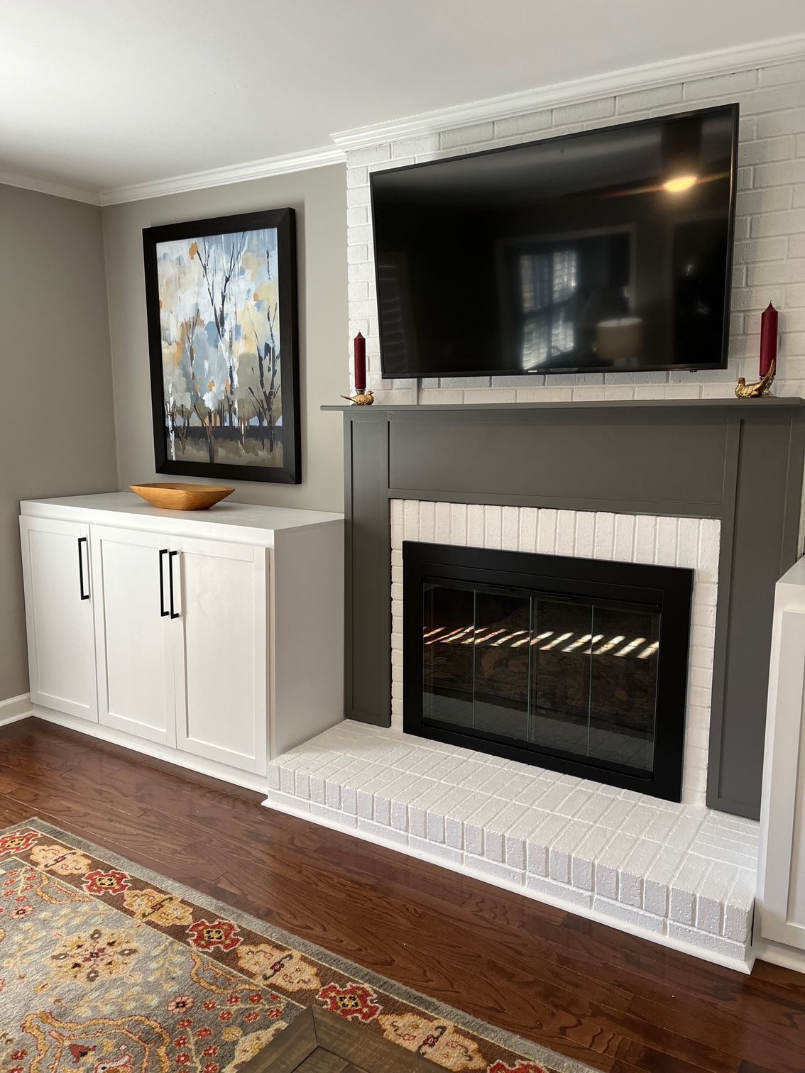 Living room with white brick fireplace, dark gray mantel, TV, and white cabinet with artwork.