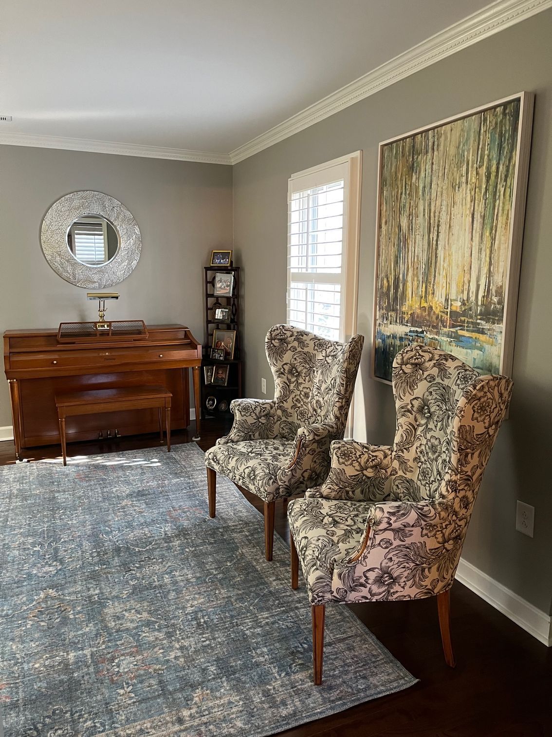 Two patterned armchairs sit near a large painting and window, across from a piano and rug.