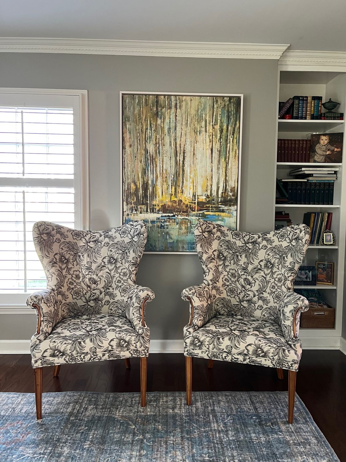 Two patterned wingback chairs face a painting above a blue rug, next to a built-in bookshelf.