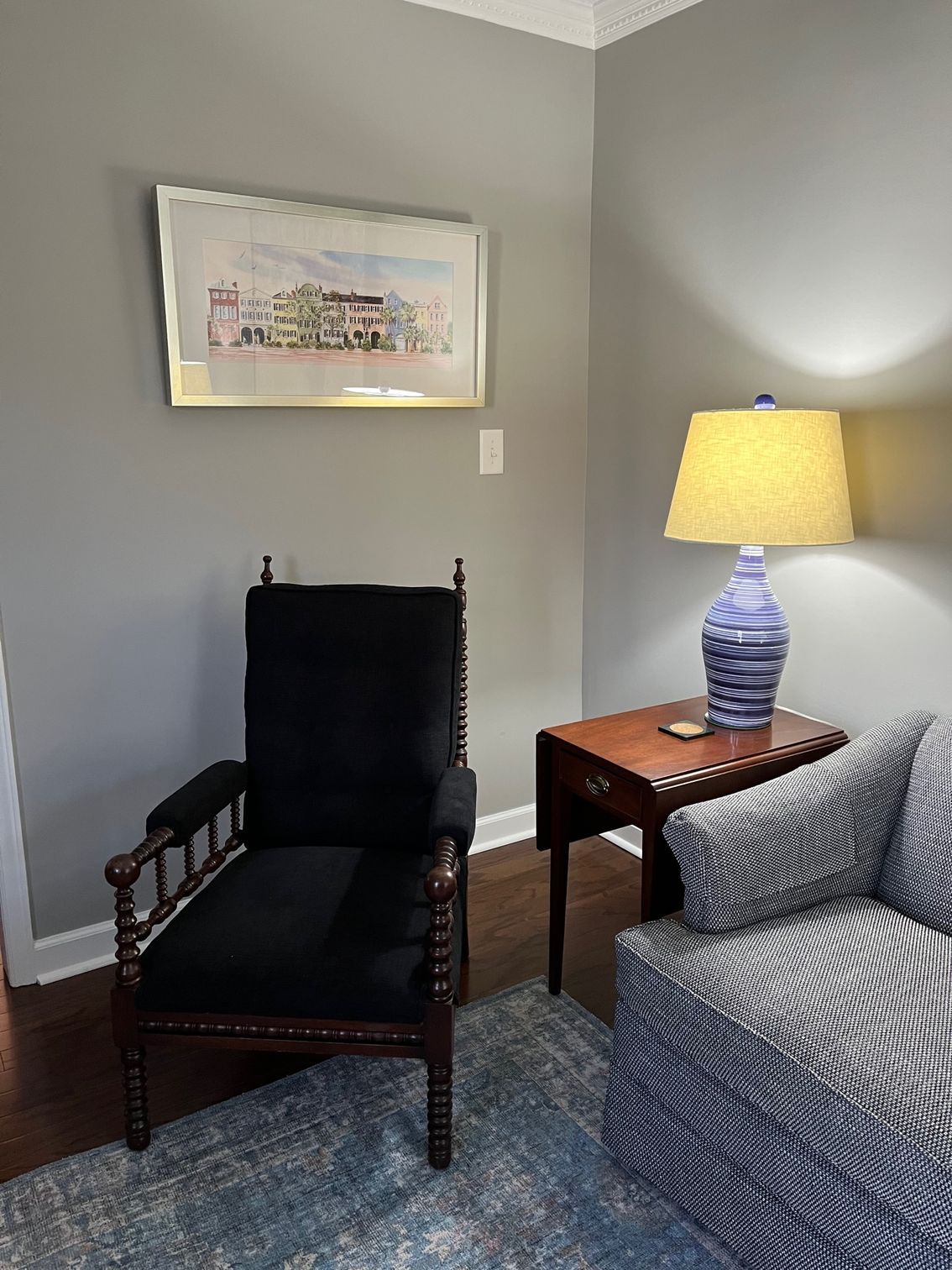 Armchair and sofa in a living room corner, with a table, lamp, and artwork on the gray wall.