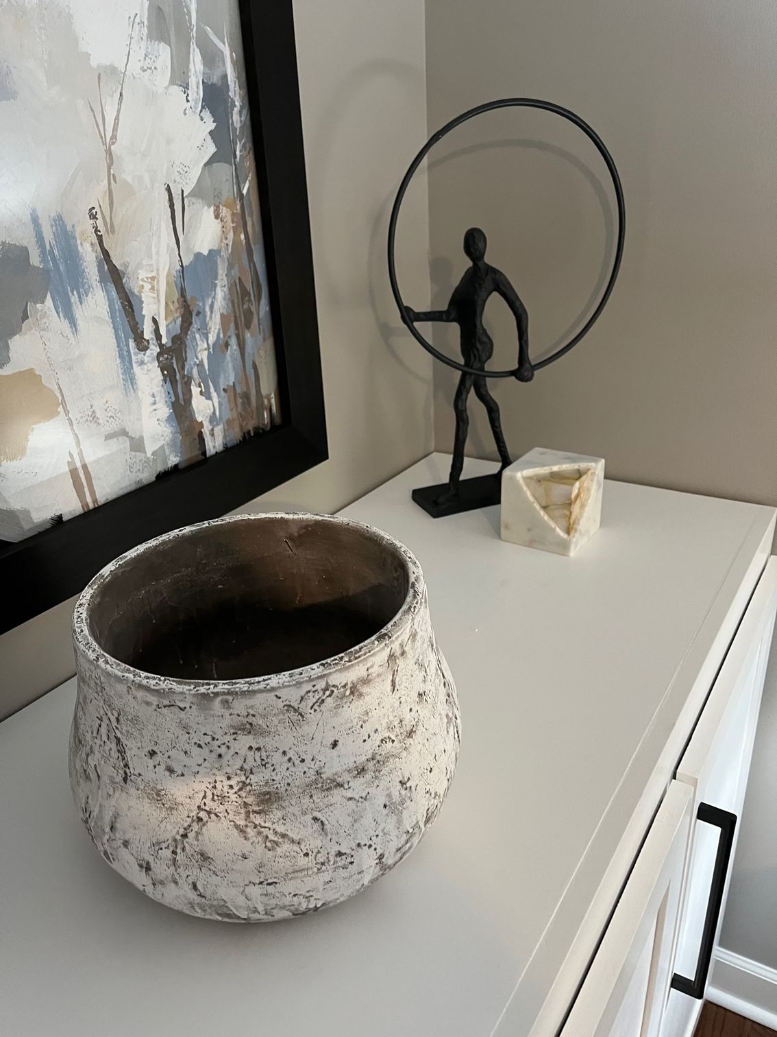 White and textured pot on a white surface next to abstract art and a black metal sculpture.