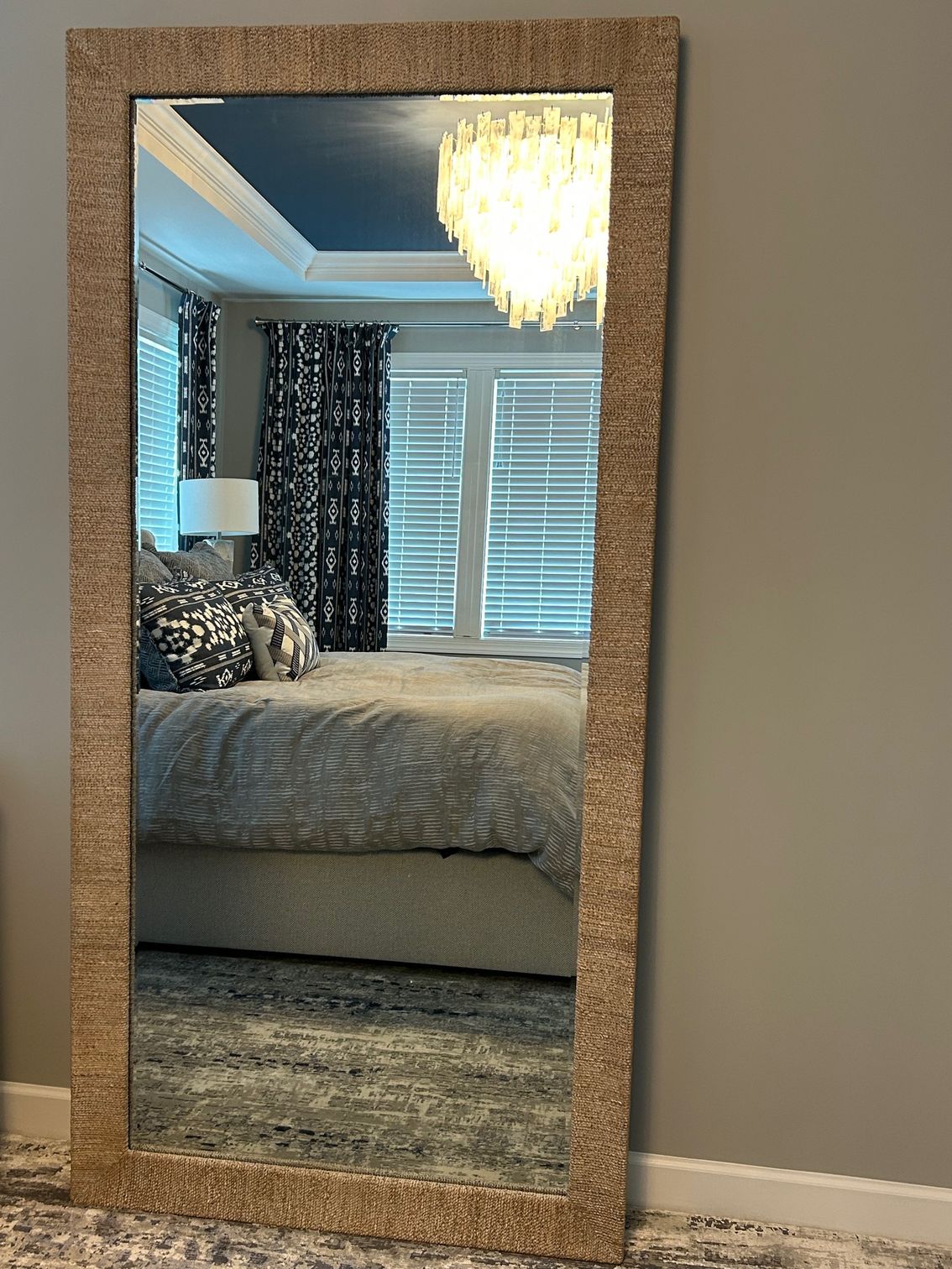 Full-length mirror with textured gold frame leans against a gray wall, reflecting a bedroom with a bed and chandelier.