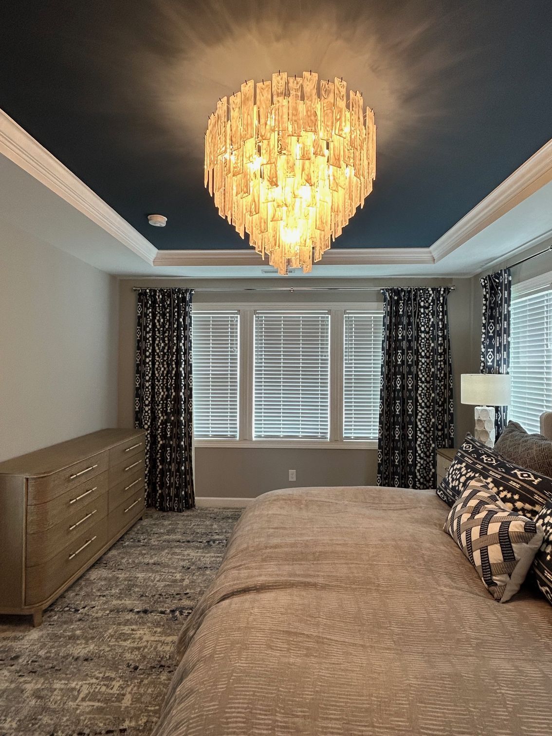 Bedroom with a large chandelier, blue ceiling, windows, patterned curtains, and neutral-colored furniture.