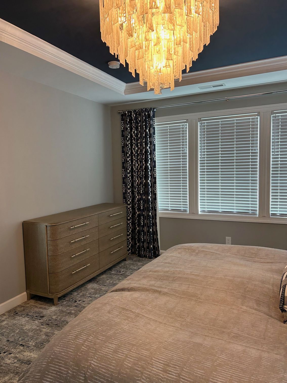 Bedroom with dresser, bed, window, patterned curtains, and ornate chandelier. Blue ceiling, neutral walls.