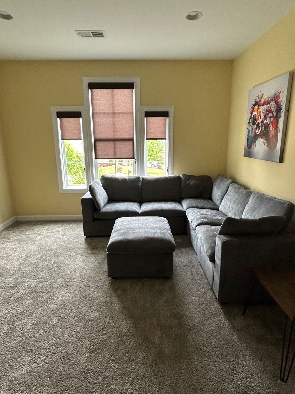 Living room with gray sectional sofa and ottoman, windows with shades, yellow walls, and a colorful painting.