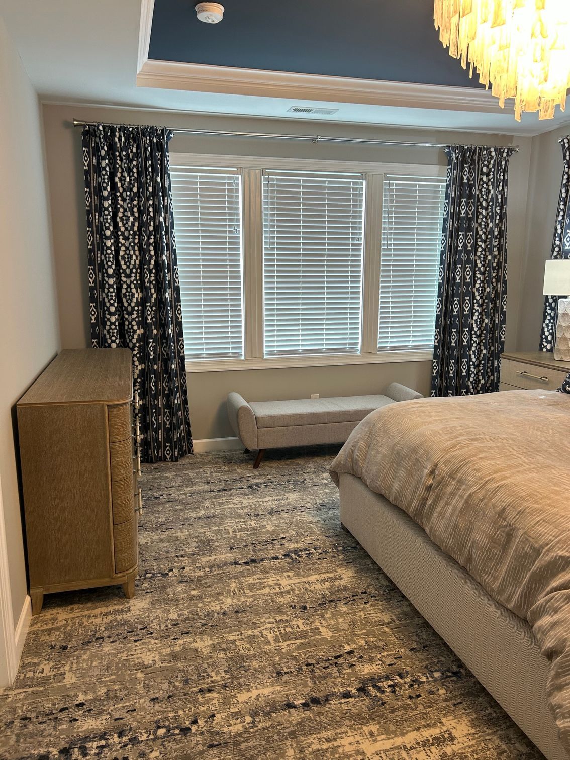 Bedroom with blue patterned curtains, a bench, and a bed. Gray rug and dresser.