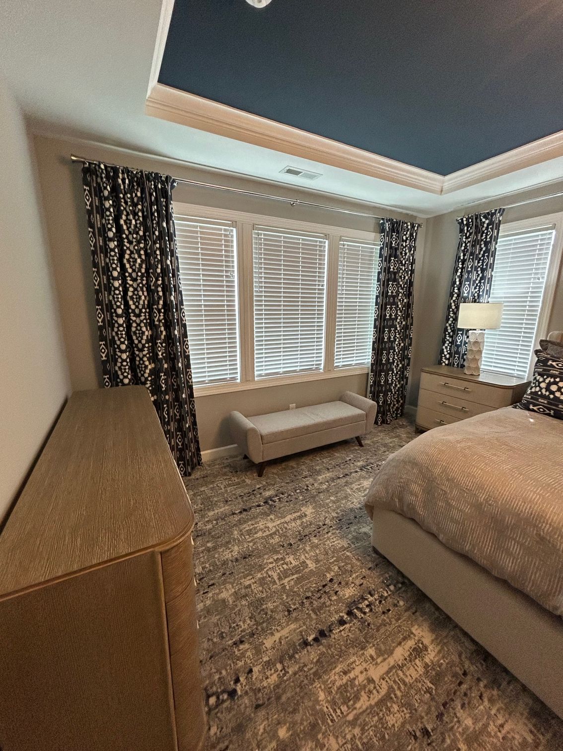 Bedroom with blue ceiling, patterned curtains, gray dresser, bench, and abstract rug.