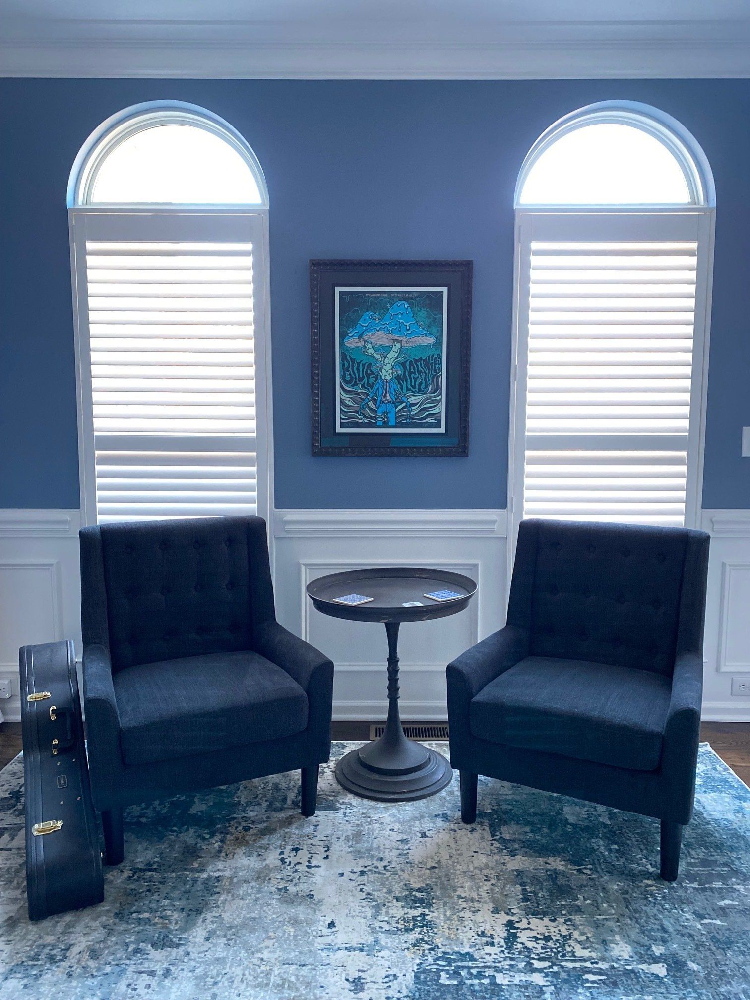 Two blue armchairs face a small table, between two arched windows. Artwork hangs on blue wall.