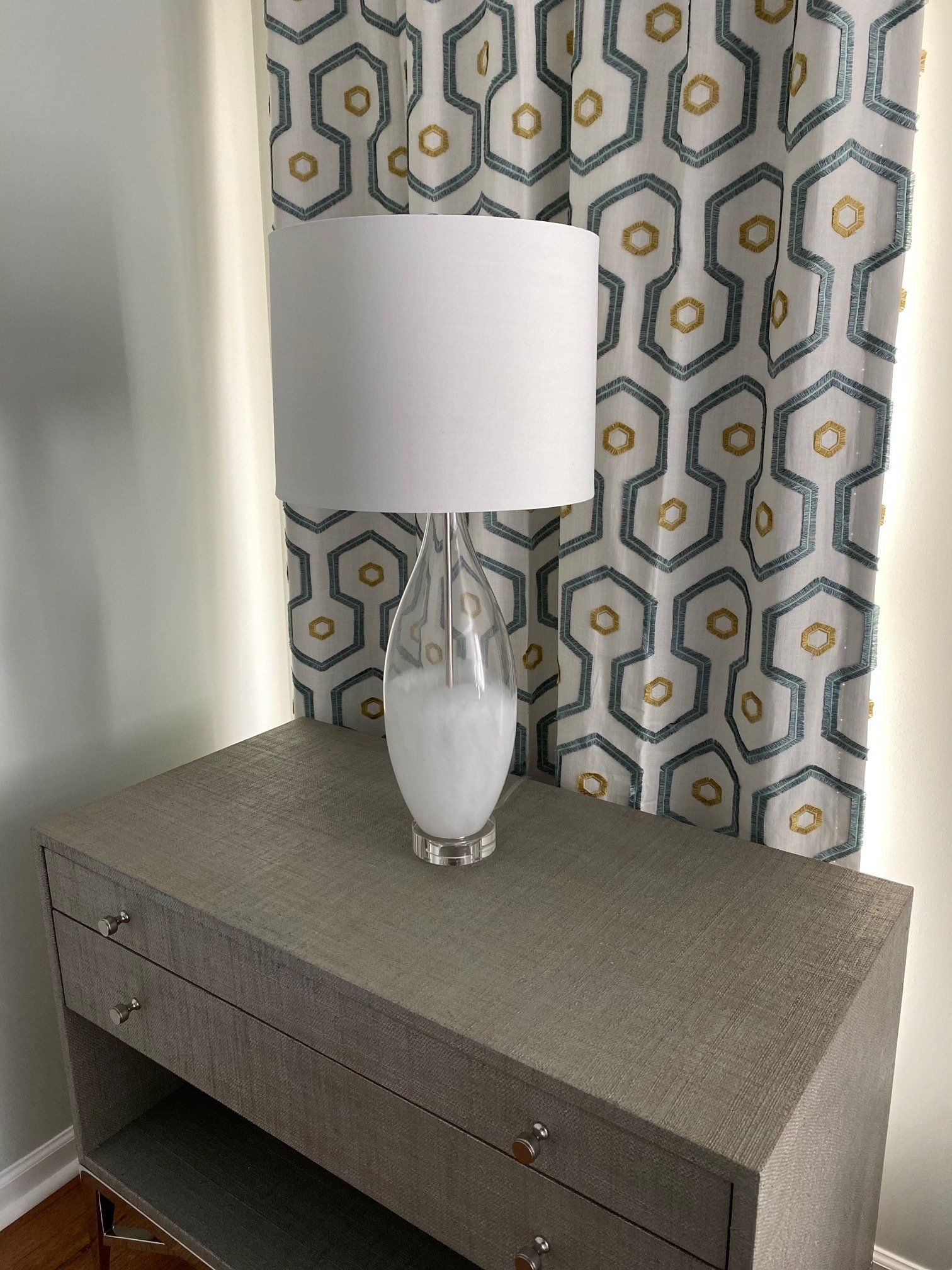 Gray textured nightstand with a white lampshade and patterned curtain.