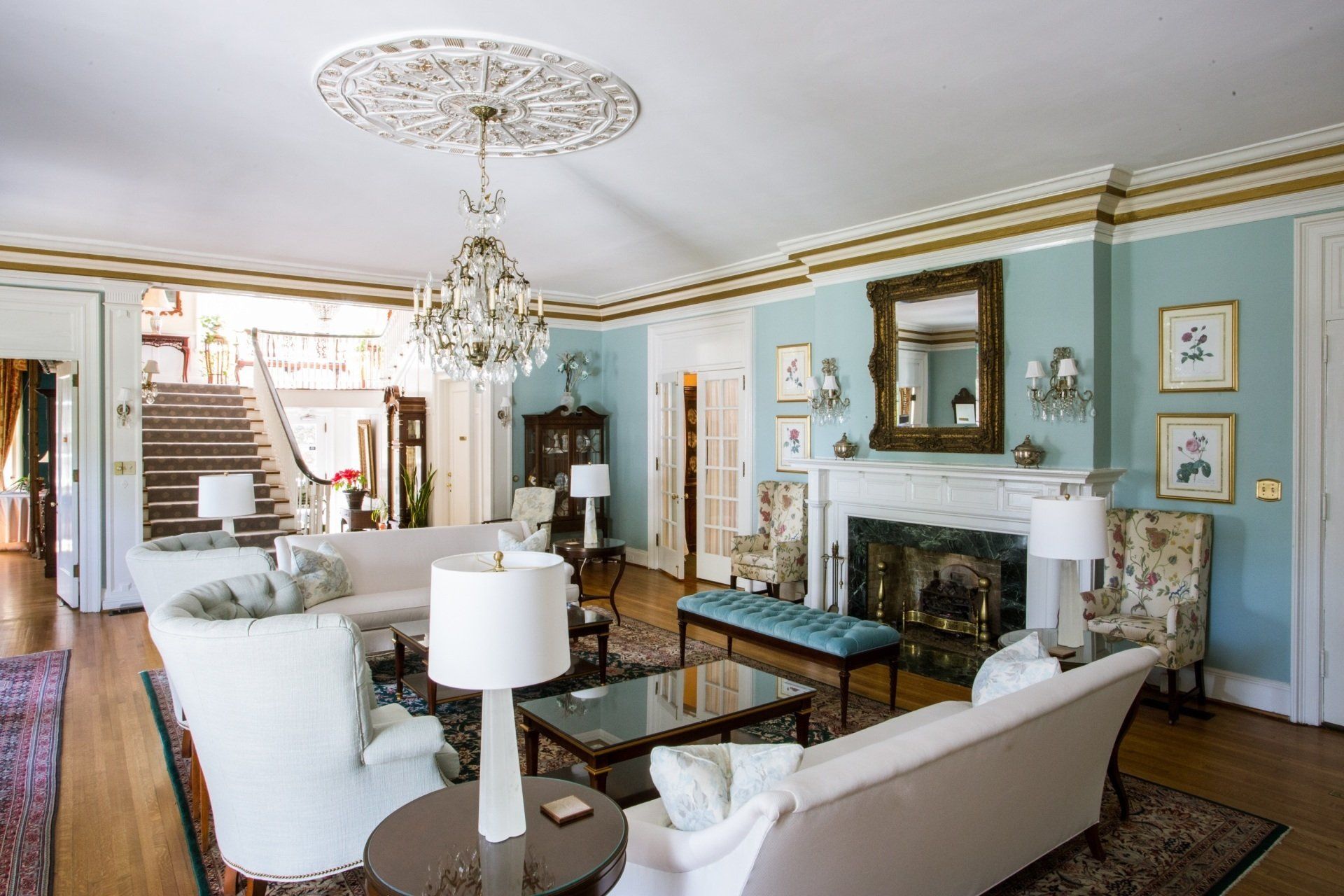 Elegant living room with blue walls, fireplace, white sofas, chandelier, and staircase.