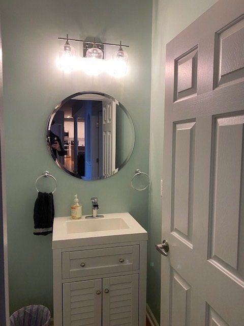 Bathroom with a white vanity, round mirror, and chrome light fixture. Light blue walls and a white door.