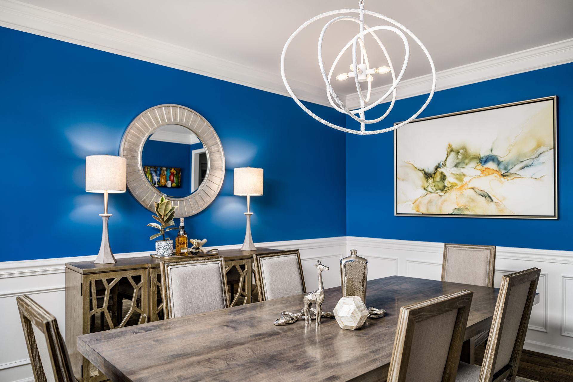 Dining room with blue walls, silver mirror, table, chairs, and modern light fixture.
