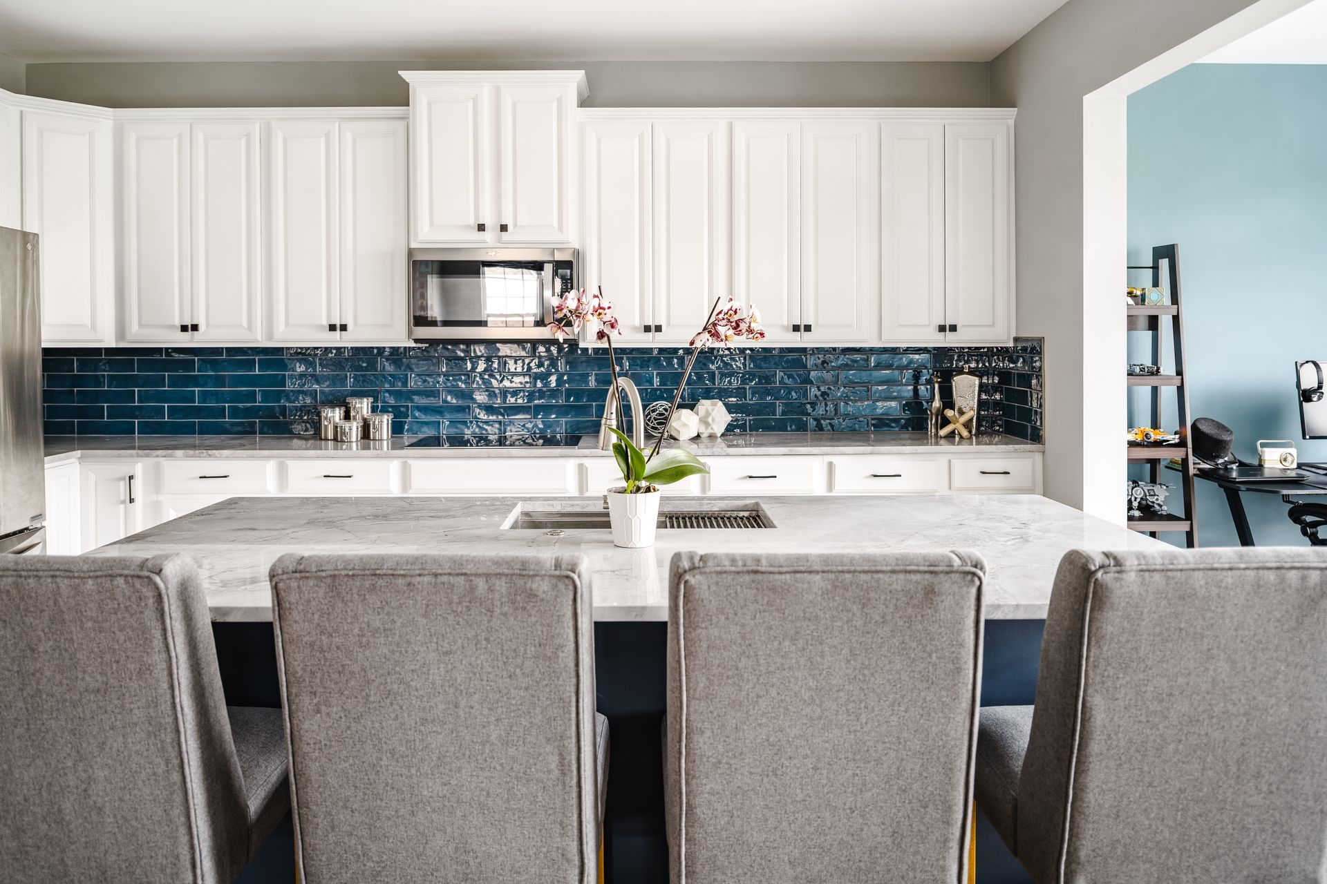 Modern kitchen with white cabinets, blue tile backsplash, gray island, and gray upholstered chairs.