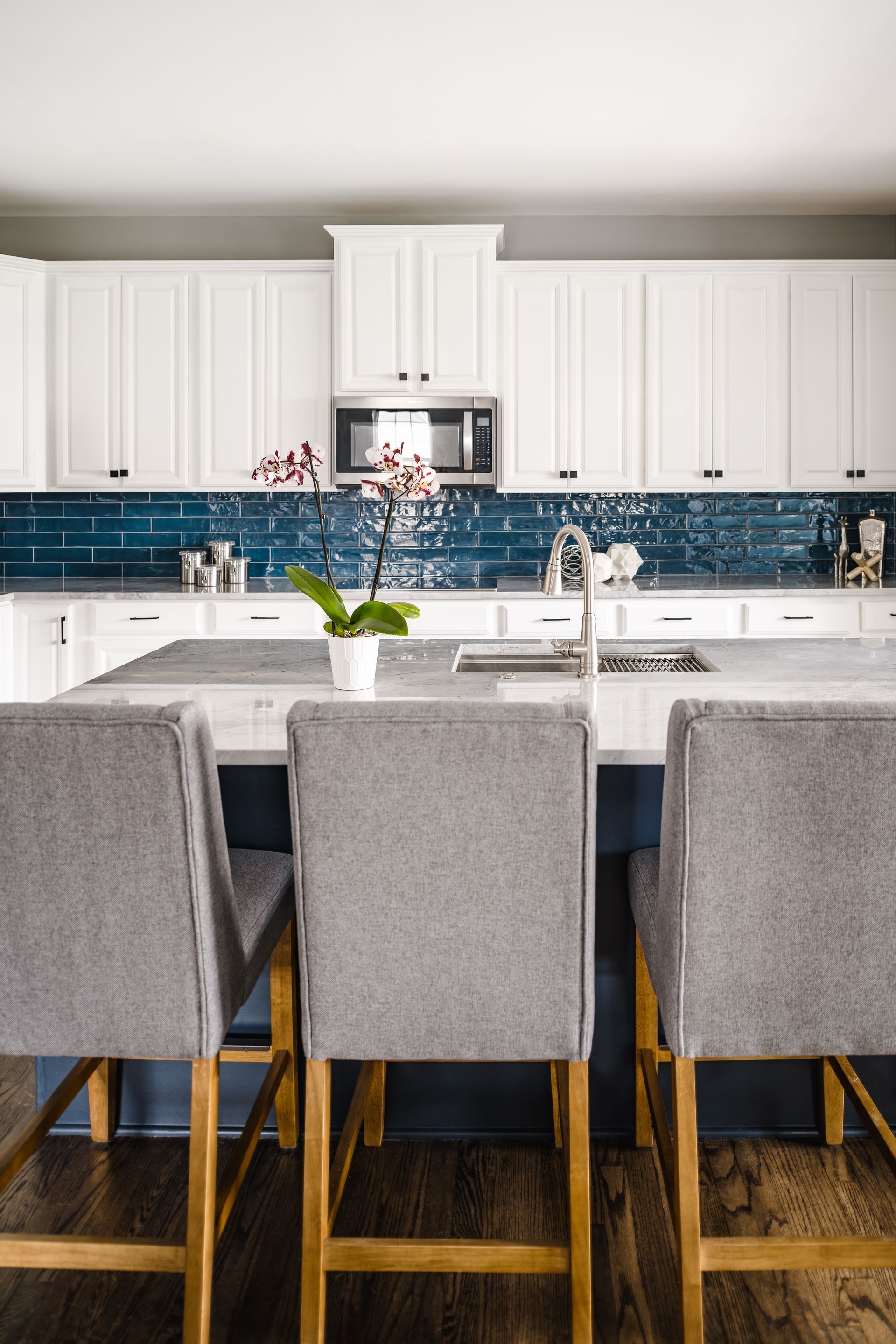 Kitchen with white cabinets, blue tile backsplash, island with grey bar stools, and an orchid.