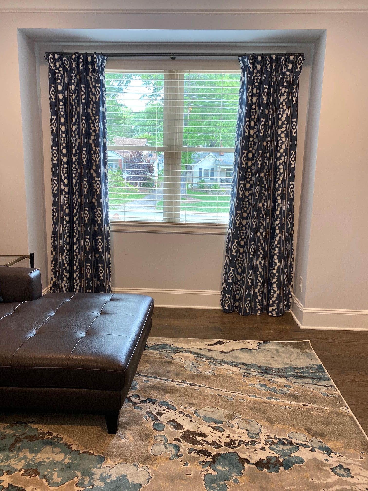 Window with patterned blue curtains and a patterned rug in a room. A leather ottoman is in the foreground.