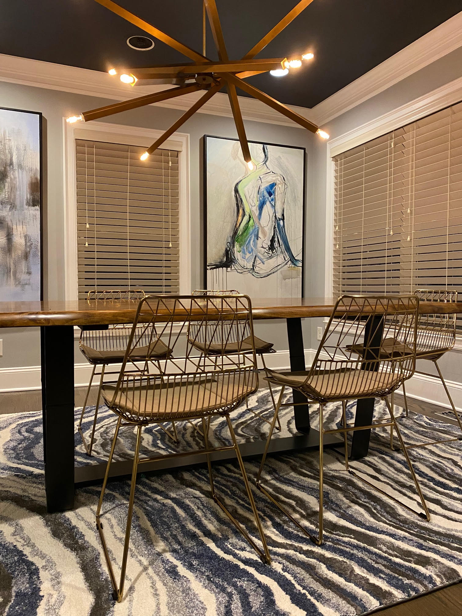 Dining room with long table, metal chairs, abstract art, and a unique chandelier. Blue and gray rug.