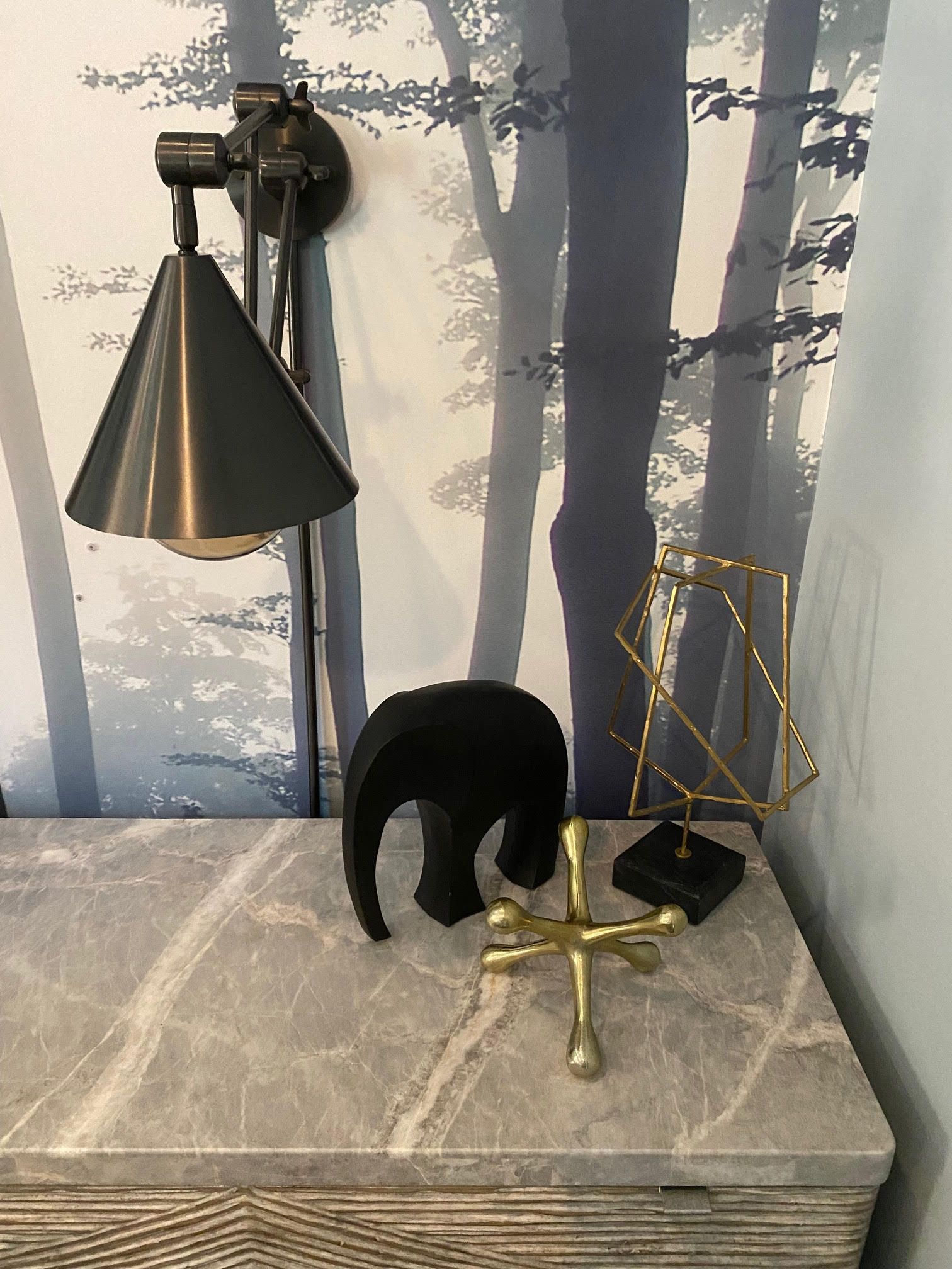 A nightstand with decor; elephant statue, gold jack, geometric sculpture, and wall lamp against a forest wallpaper.