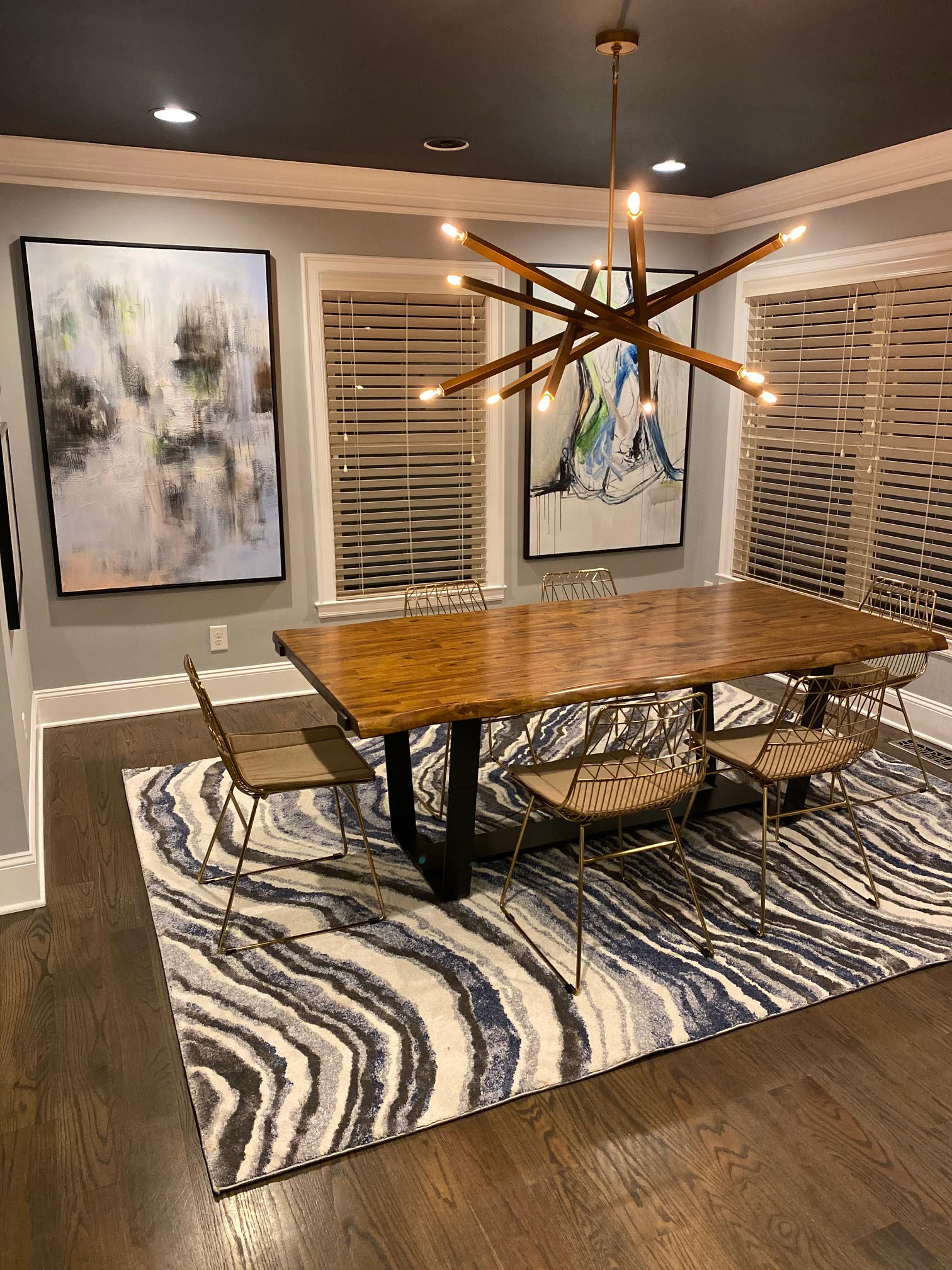 Dining room with long wooden table, art, and patterned rug; dark ceiling and modern chandelier.