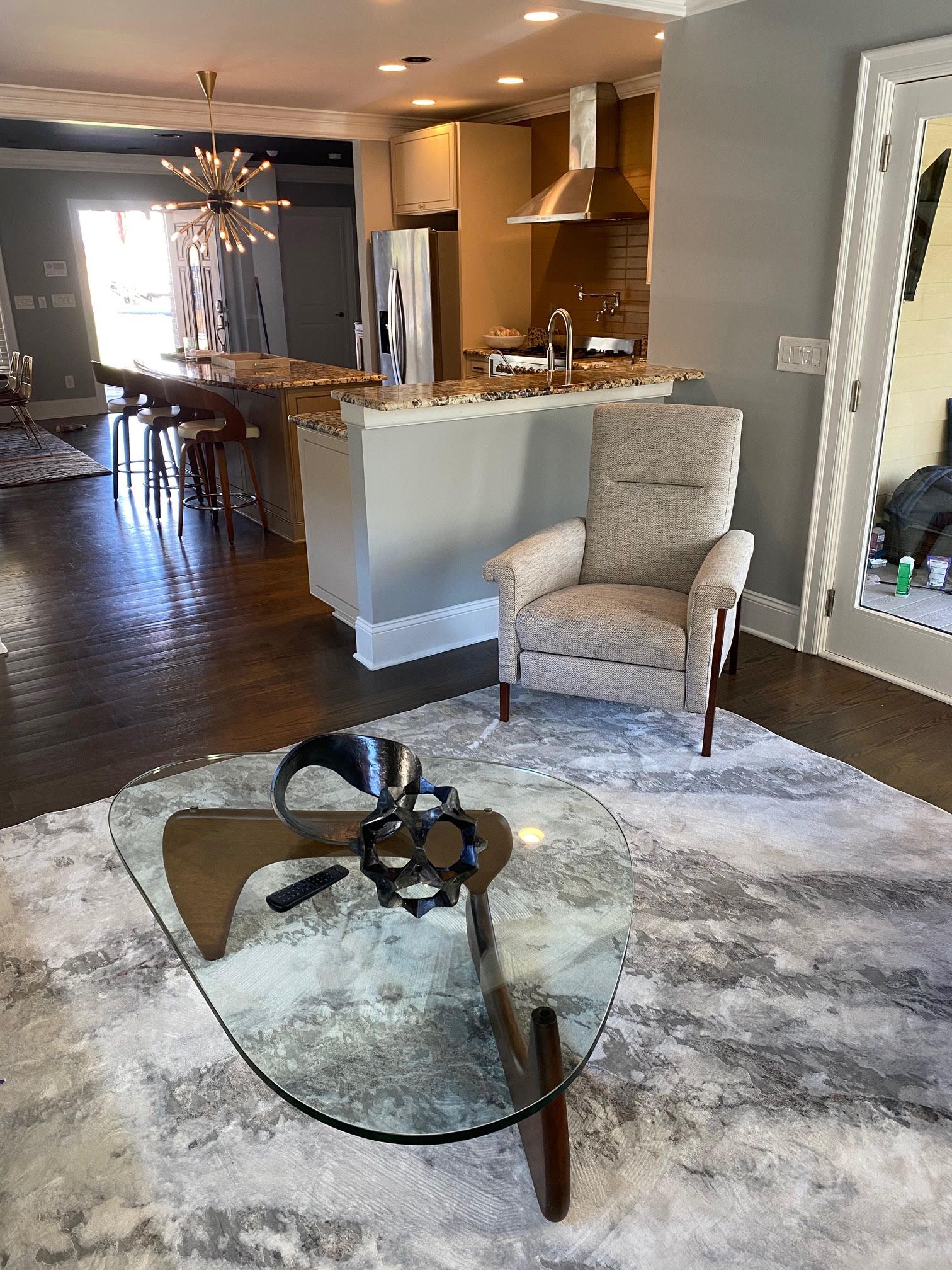 Living room with patterned rug, unique coffee table, beige armchair, and kitchen in the background.