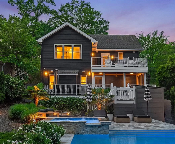 Dark gray two-story house with balconies and pool. Lush landscaping, trees in the background.