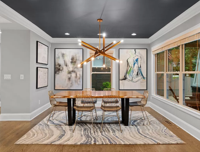 Modern dining room with wood table, abstract art, and gold light fixture. Gray walls, dark ceiling, and area rug.