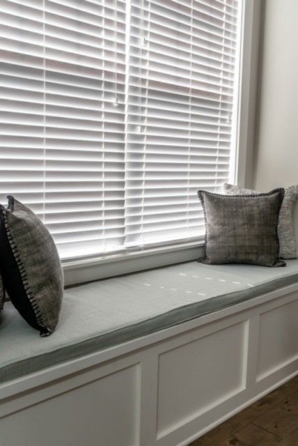 White window seat with gray cushions and pillows; white blinds in a sunny room.