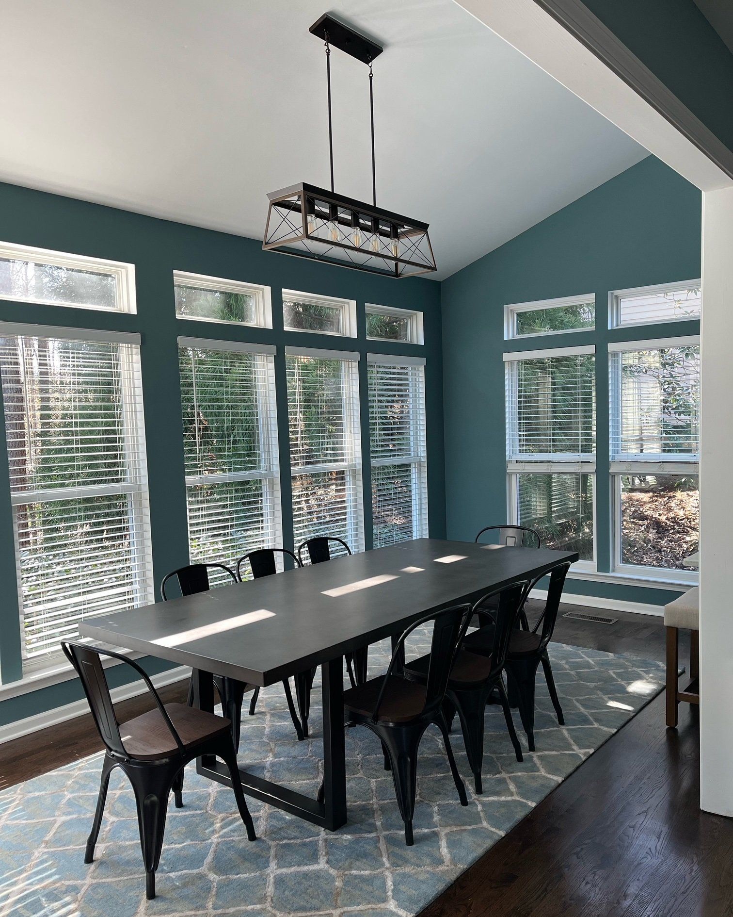 Dining room with a long black table, black metal chairs, and a teal accent wall with large windows.