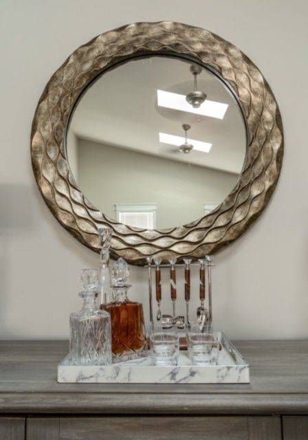 Bar setup with liquor decanters, glasses, and a round mirror on a gray cabinet.