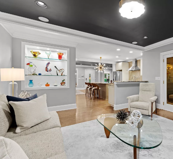 Living room with open kitchen, light gray walls, dark gray ceiling, glass coffee table, and glass-shelved display case.