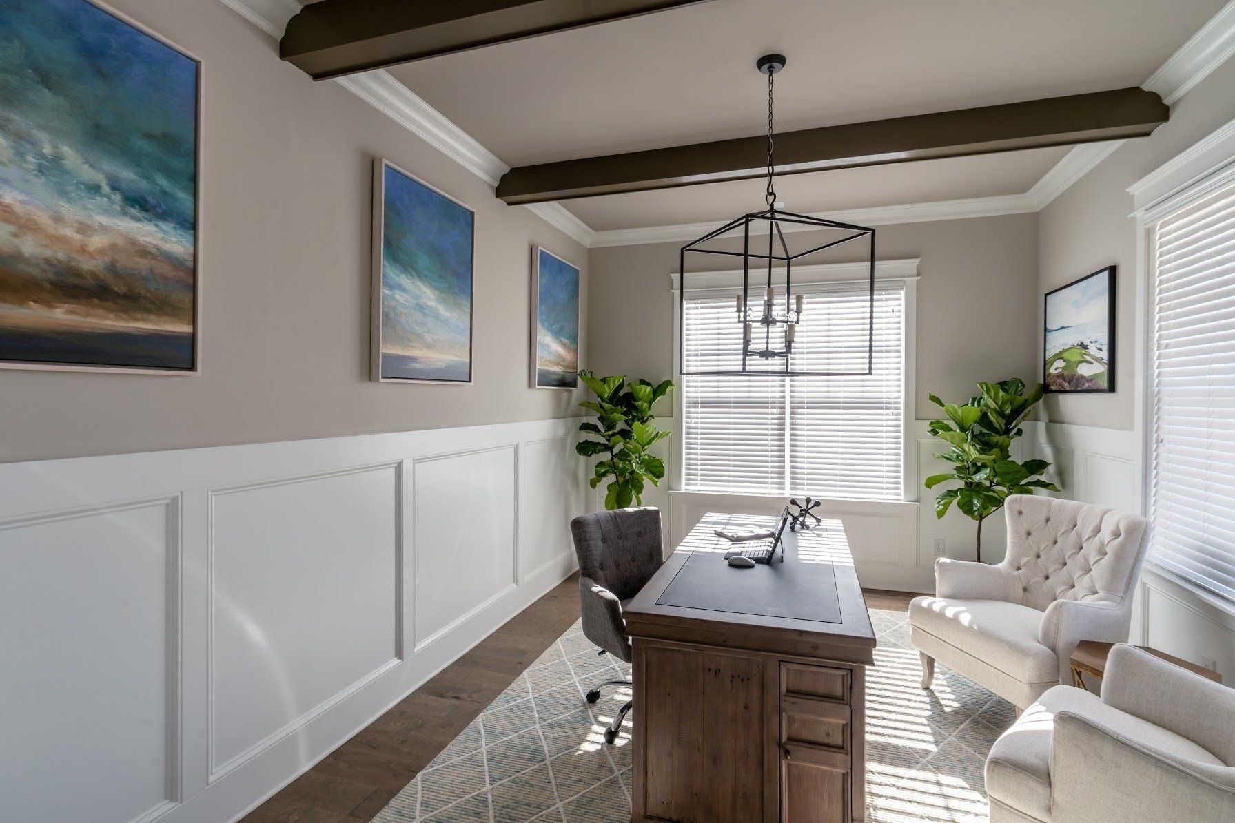 Home office with art, wood desk, tufted chairs, and large windows with blinds.