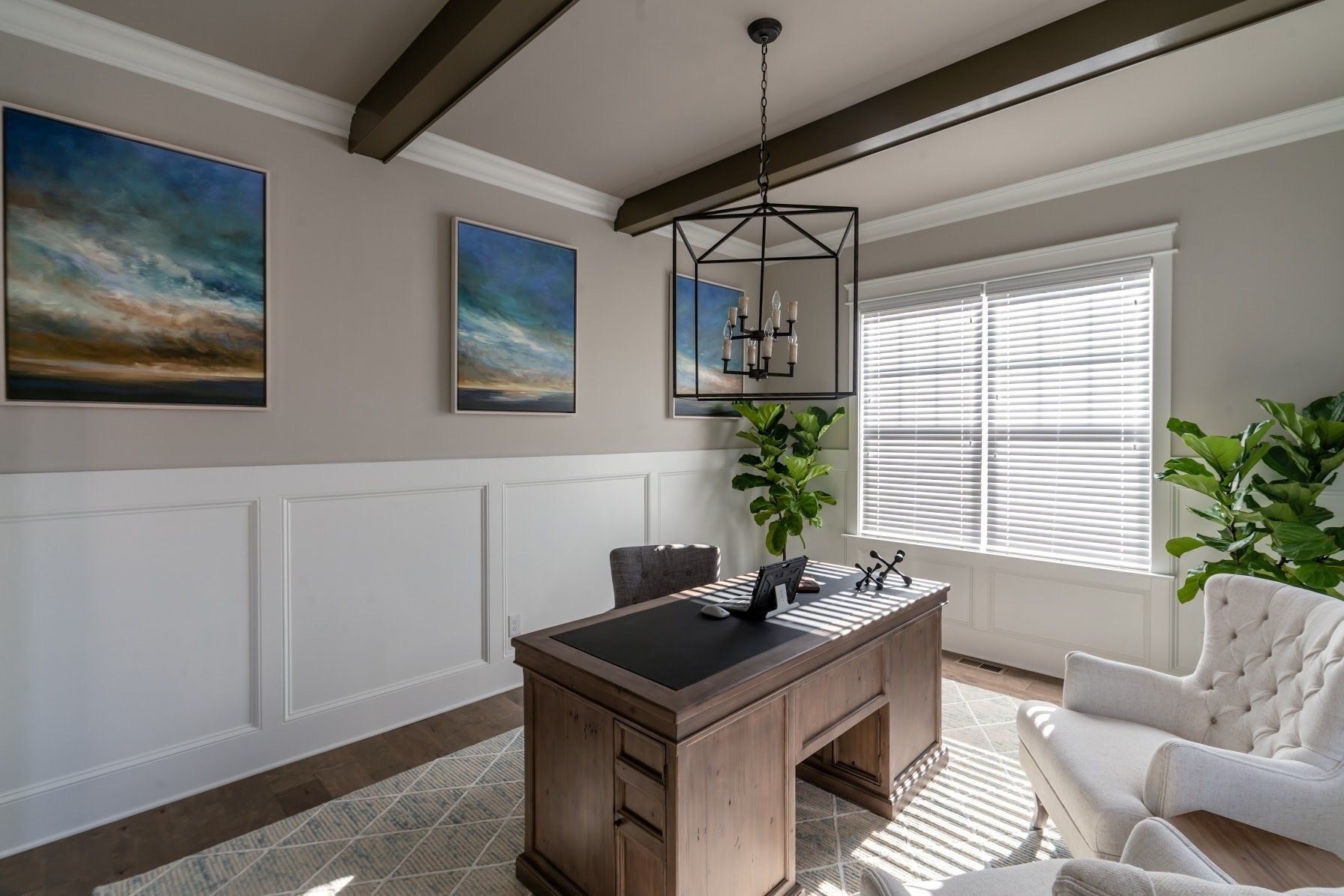 Home office with desk, art, two white armchairs, and plants, and ceiling beams.