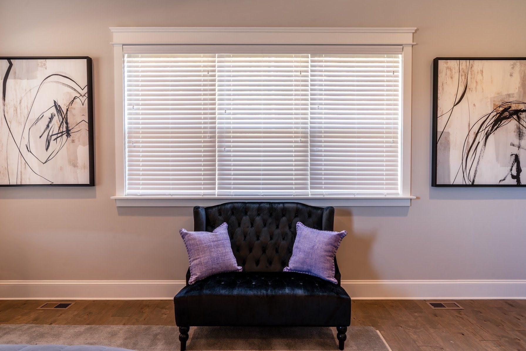 Black chair with purple pillows beneath a window with white blinds, flanked by abstract art.