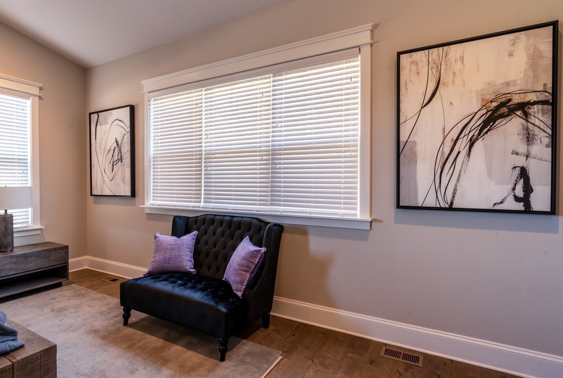Room with a black tufted armchair, two art pieces, and a window with blinds; beige walls and carpet.