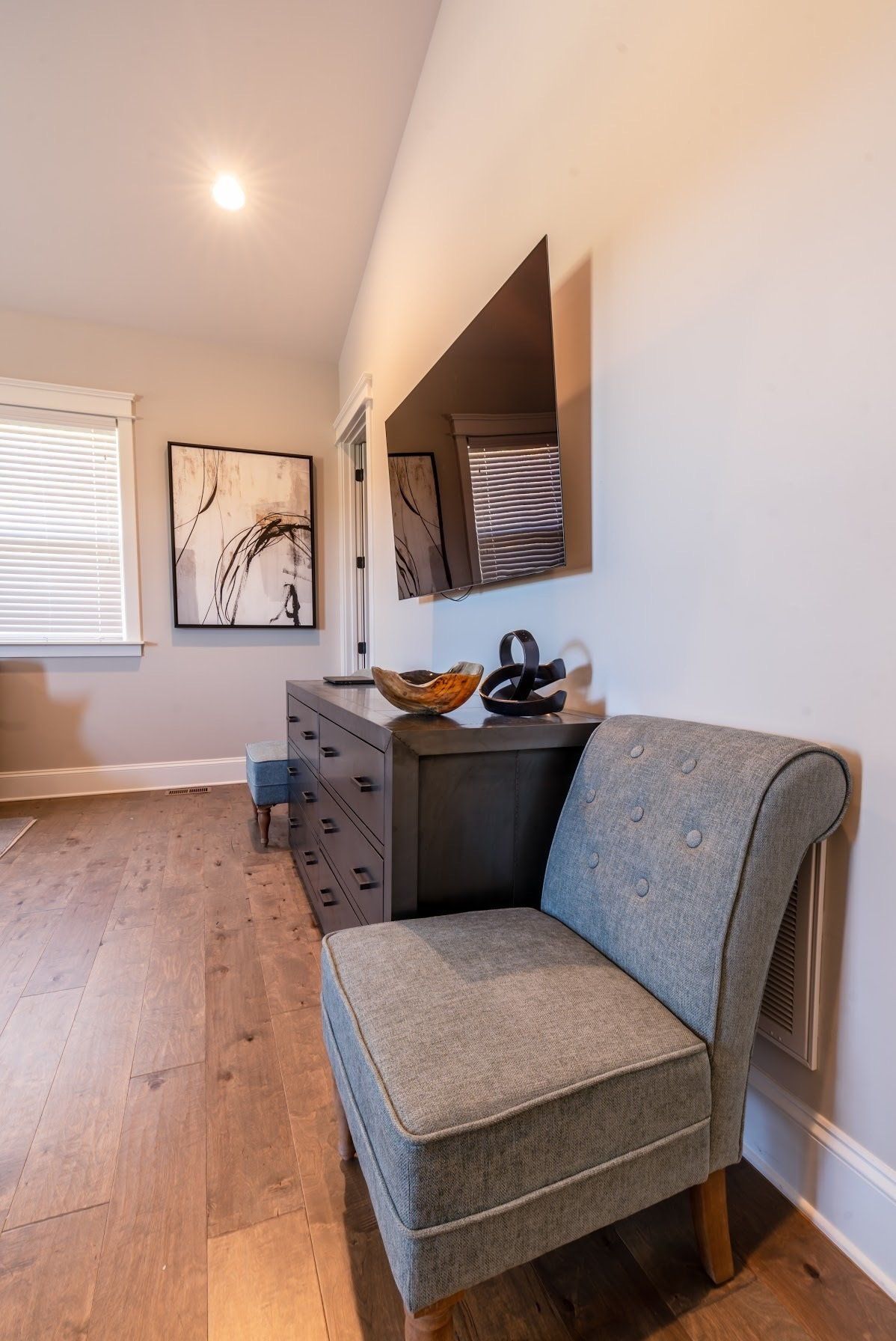 A bedroom with hardwood floors, a dresser, and a chair. A TV is mounted on the wall.