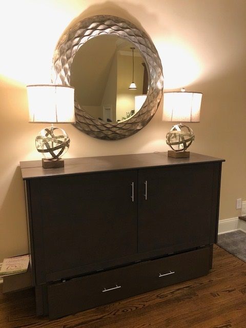 Dark brown cabinet with two lamps, a circular mirror, and a drawer.