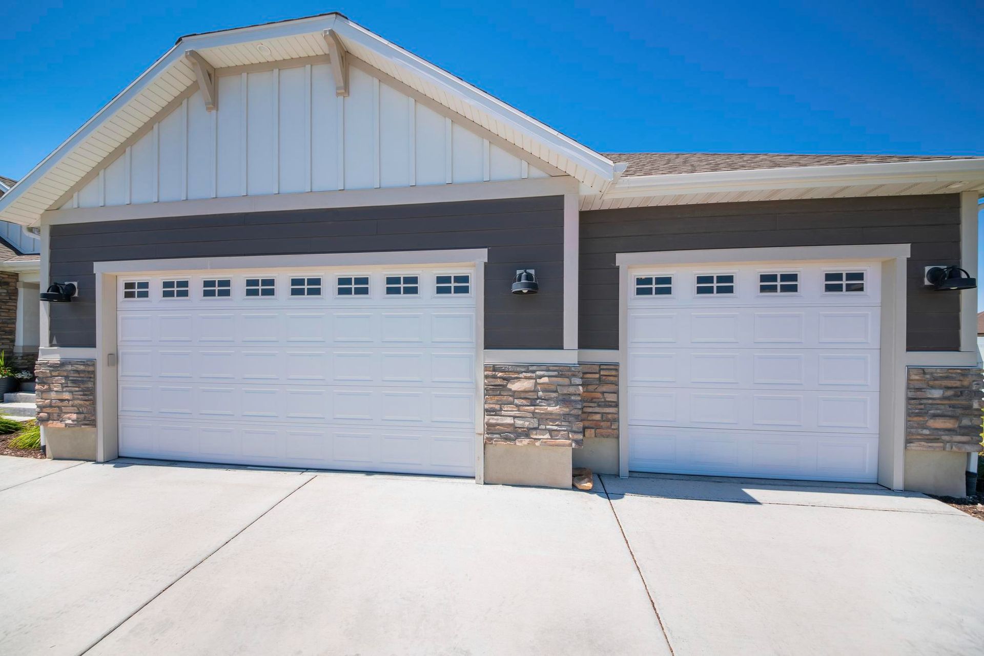 Two-car garage with white doors, gray siding, stone accents, and a concrete driveway on a sunny day.