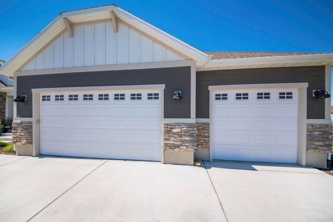 Two-car garage with white doors, gray siding, stone accents, and a concrete driveway on a sunny day.